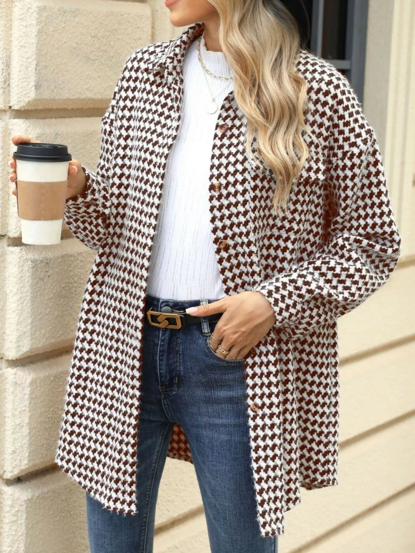 Woman in houndstooth button-up long sleeve coat, holding coffee, styled casually with jeans.