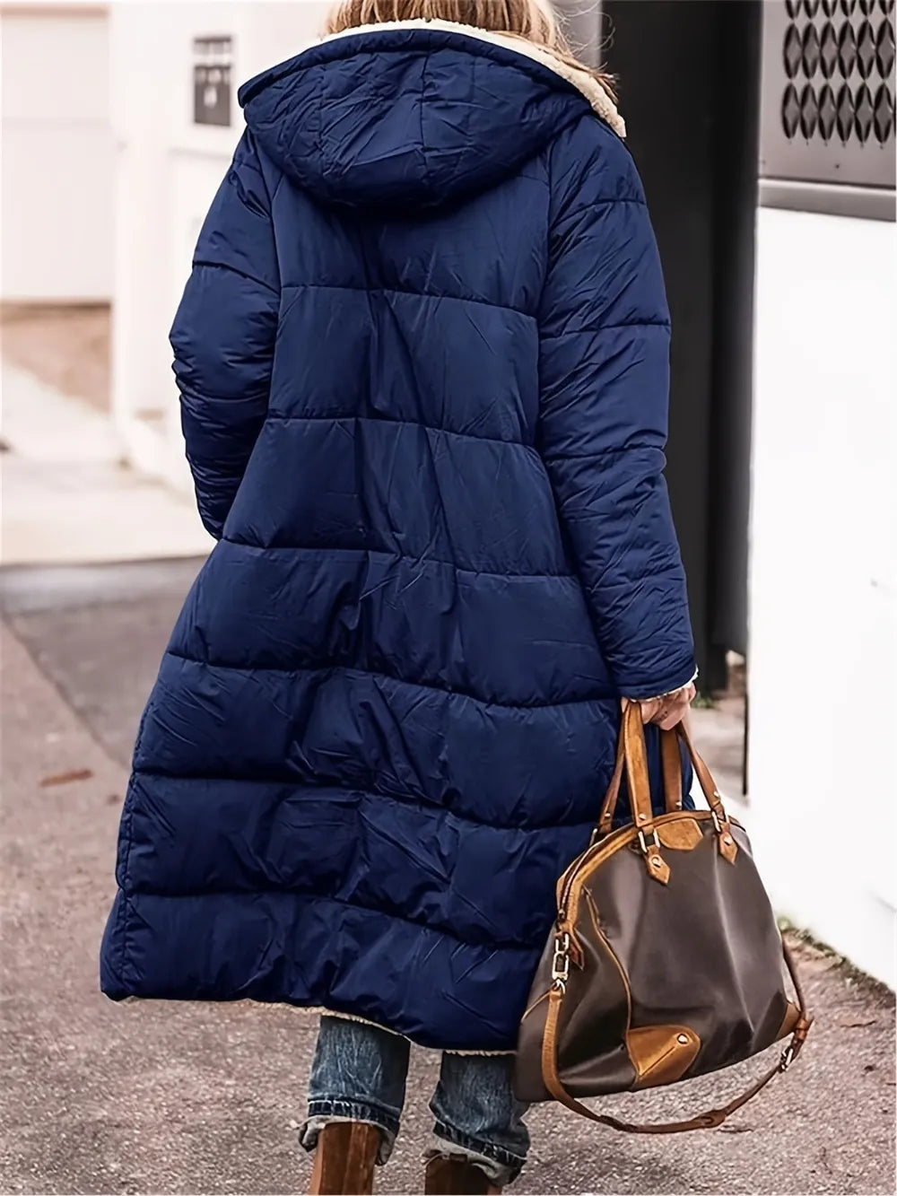 Model wearing a full size navy zip-up Sherpa hooded coat, carrying a brown handbag, walking on a city street.