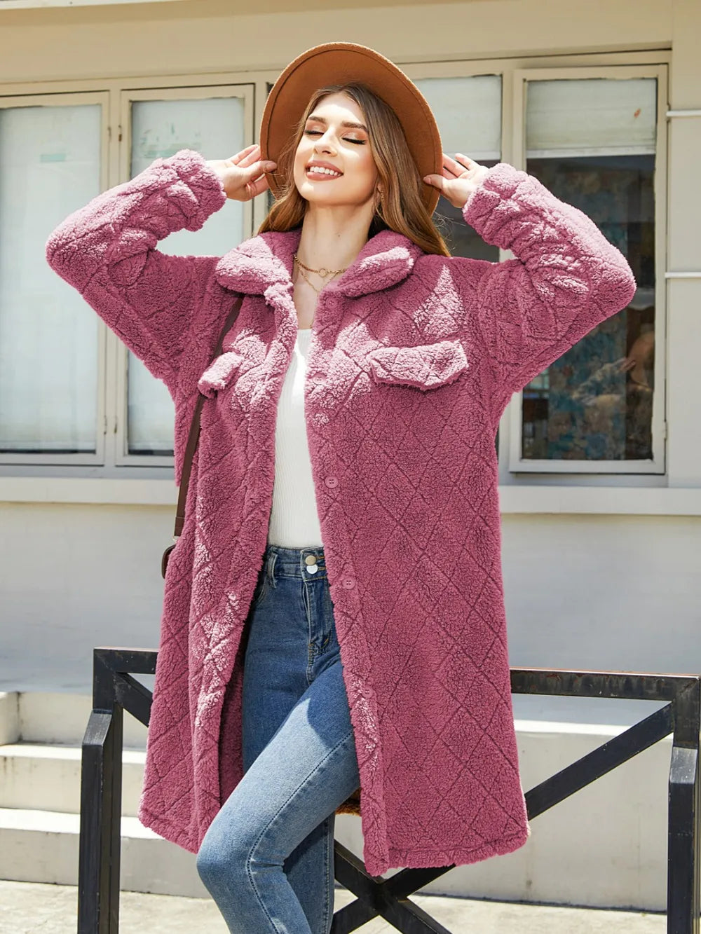 Woman enjoying the sun in a pink textured button-up coat with pockets and a floppy hat.